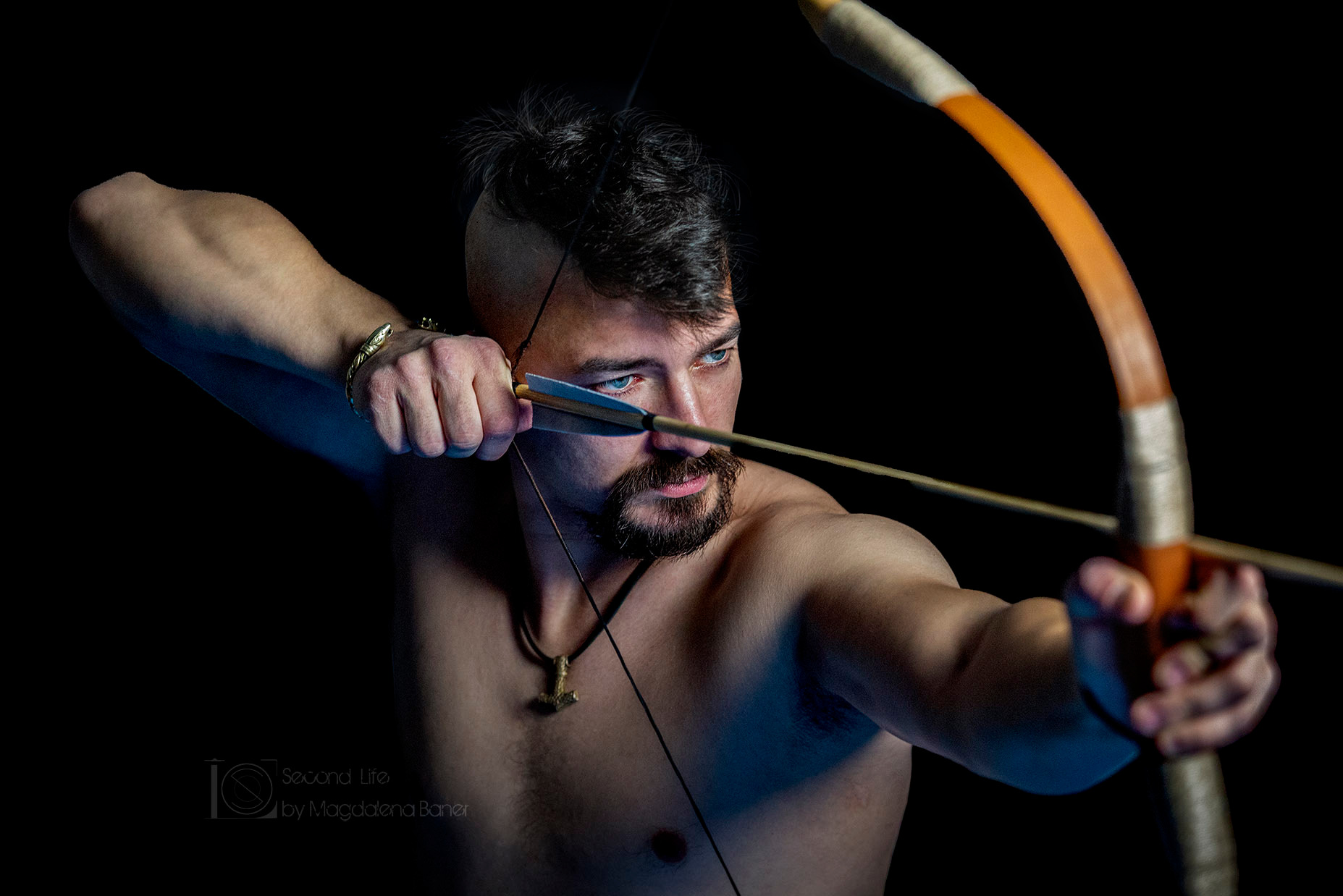 Mężczyzna z irokezem i brodą, naciągający łuk, z tatuażem na ramieniu, w złotej bransoletce w kształcie węża, na czarnym tle, z widocznym logo fotografa w lewym dolnym rogu.