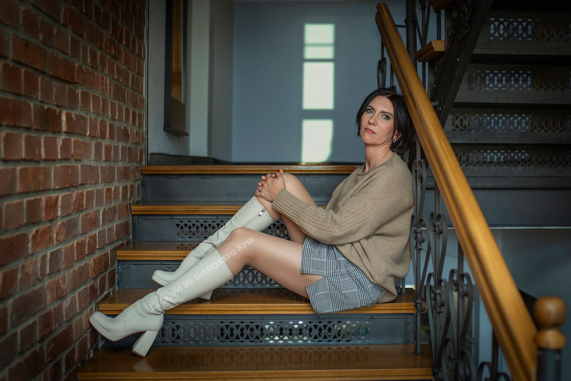 Kobieta w beżowym swetrze i białych kozakach siedzi na drewnianych schodach z metalową balustradą, ceglana ściana w tle.
