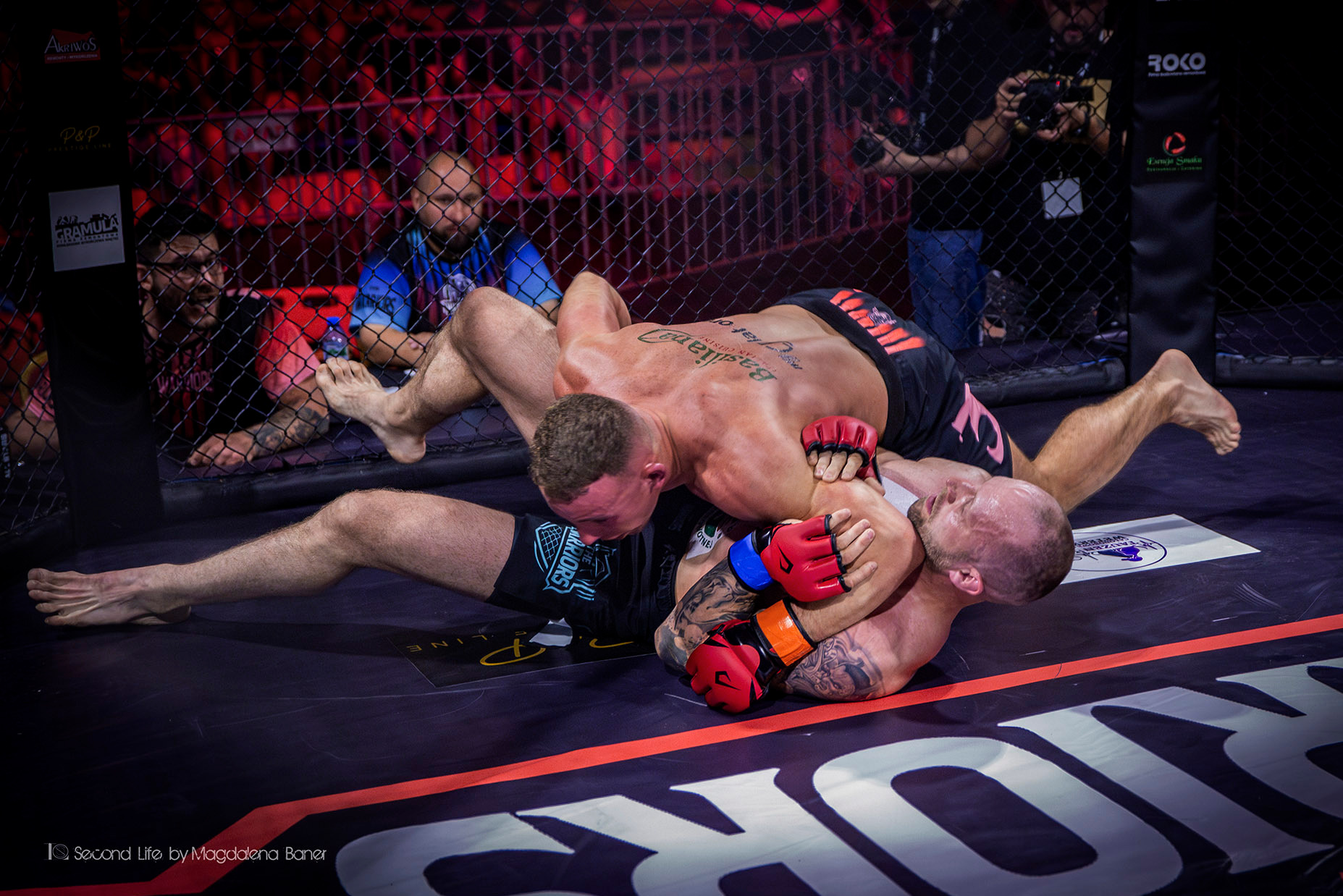 Dwóch zawodników MMA w parterze podczas walki w klatce, jeden z nich w czarnych spodenkach z napisem 'The Warriors', drugi z tatuażem na plecach, obaj w czerwonych rękawicach.