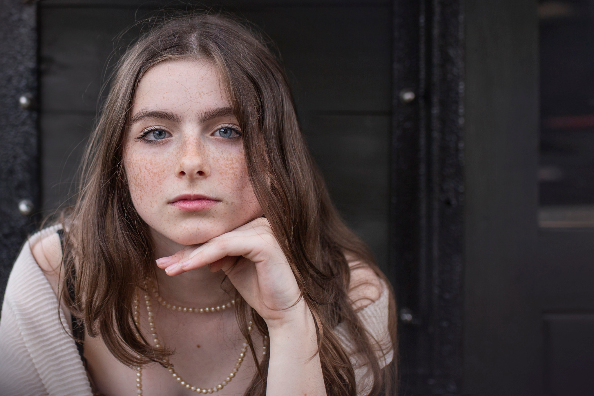 Portret młodej kobiety z piegami, opierającej podbródek na dłoni, w jasnej bluzce i perłowym naszyjniku, na tle ciemnych drzwi.
