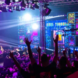 Widok na scenę podczas gali wrestlingu z publicznością, intensywne fioletowe oświetlenie, ekrany LED z grafikami i logotypami, stalowa konstrukcja podtrzymująca oświetlenie.