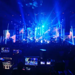 Koncert w Spodku: realizacja sygnału wideo na ekrany, widoczny mikser wideo i ekrany podglądowe, liczne reflektory sceniczne w kolorze niebieskim, publiczność w tle.