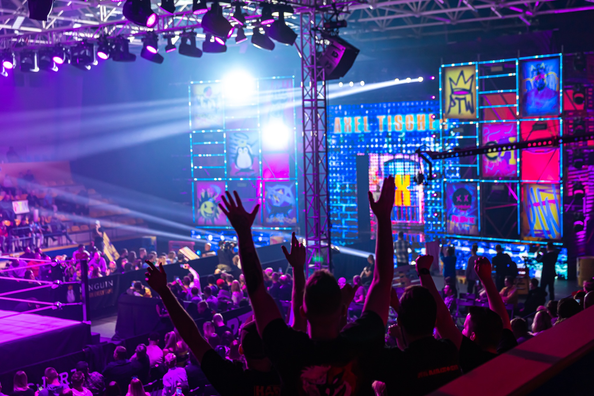 Widok na scenę podczas gali wrestlingu z publicznością, intensywne fioletowe oświetlenie, ekrany LED z grafikami i logotypami, stalowa konstrukcja podtrzymująca oświetlenie.