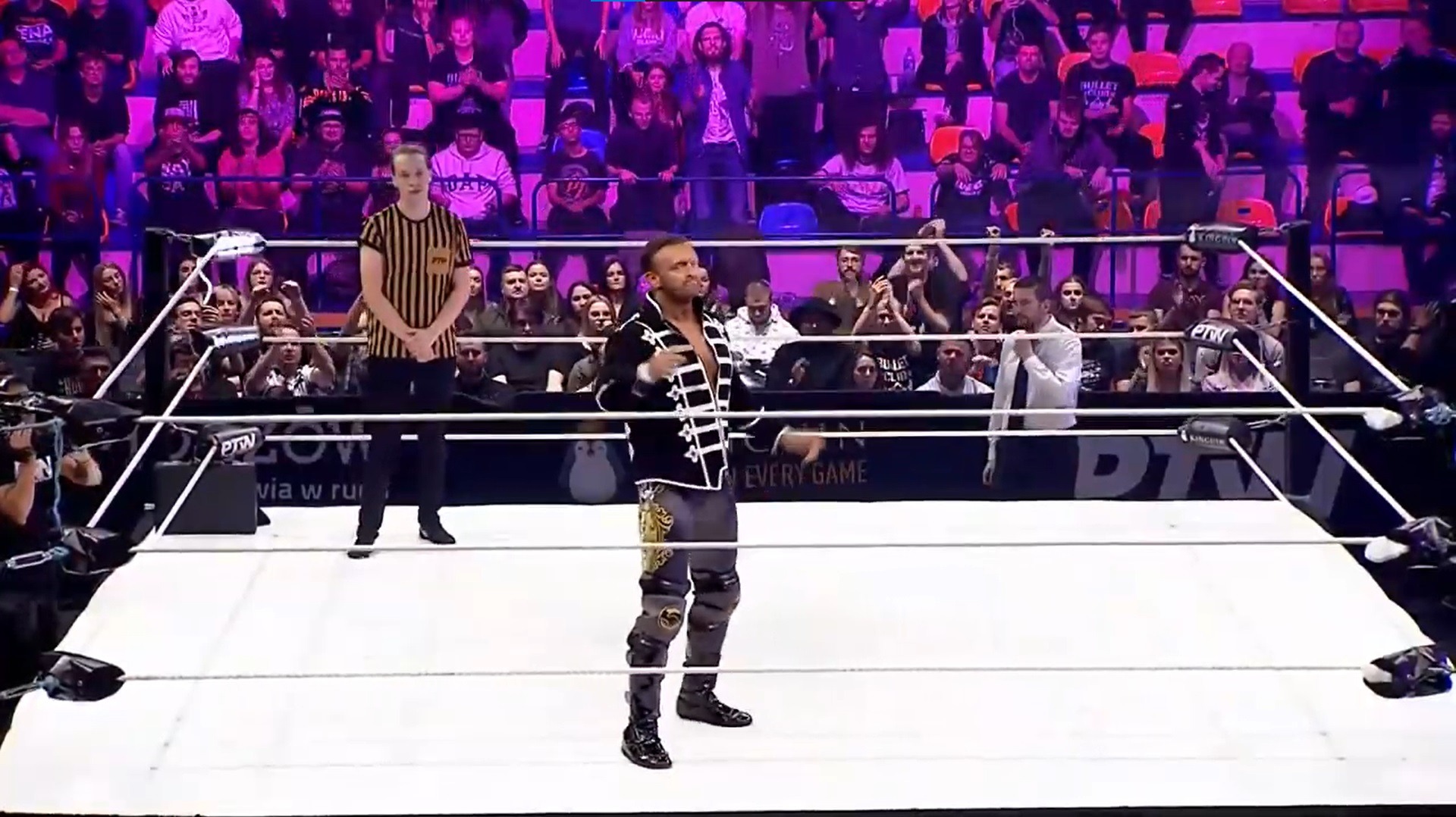Mężczyzna w ringu w stroju z czarną marynarką w stylu wojskowym i ozdobnymi spodniami, sędzia w tle, publiczność na trybunach podczas gali wrestlingu Prime Time Wrestling.
