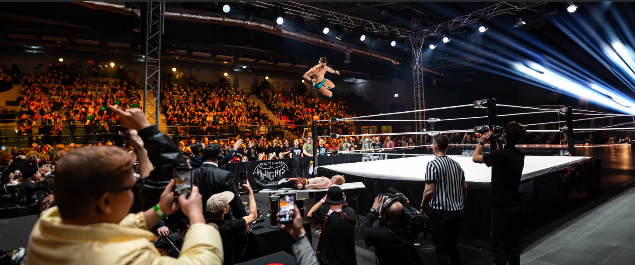 Dynamiczne ujęcie z gali wrestlingu: zawodnik w locie nad ringiem, oświetlenie sceniczne, publiczność w tle, filmowcy rejestrujący akcję.