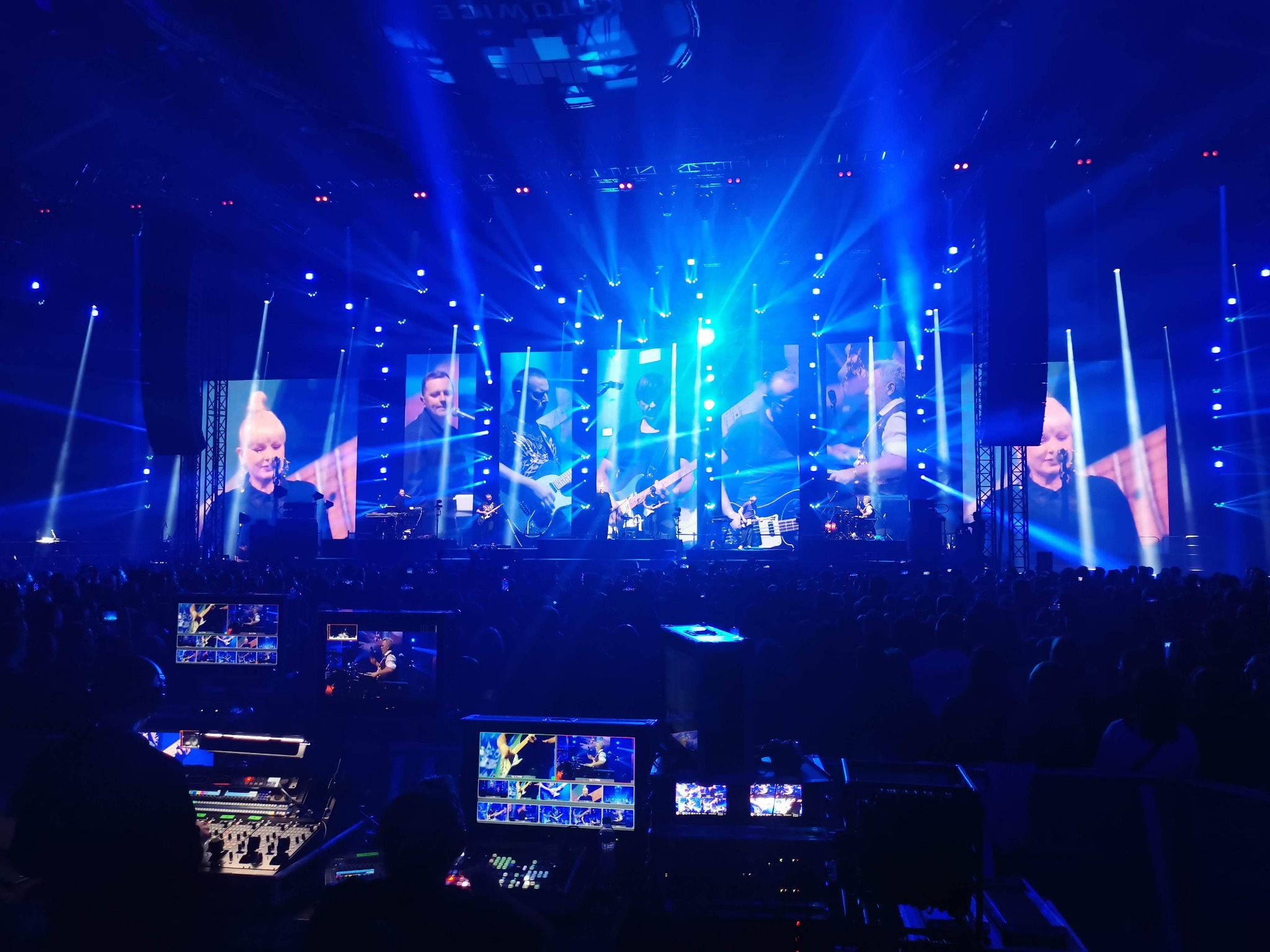 Koncert w Spodku: realizacja sygnału wideo na ekrany, widoczny mikser wideo i ekrany podglądowe, liczne reflektory sceniczne w kolorze niebieskim, publiczność w tle.
