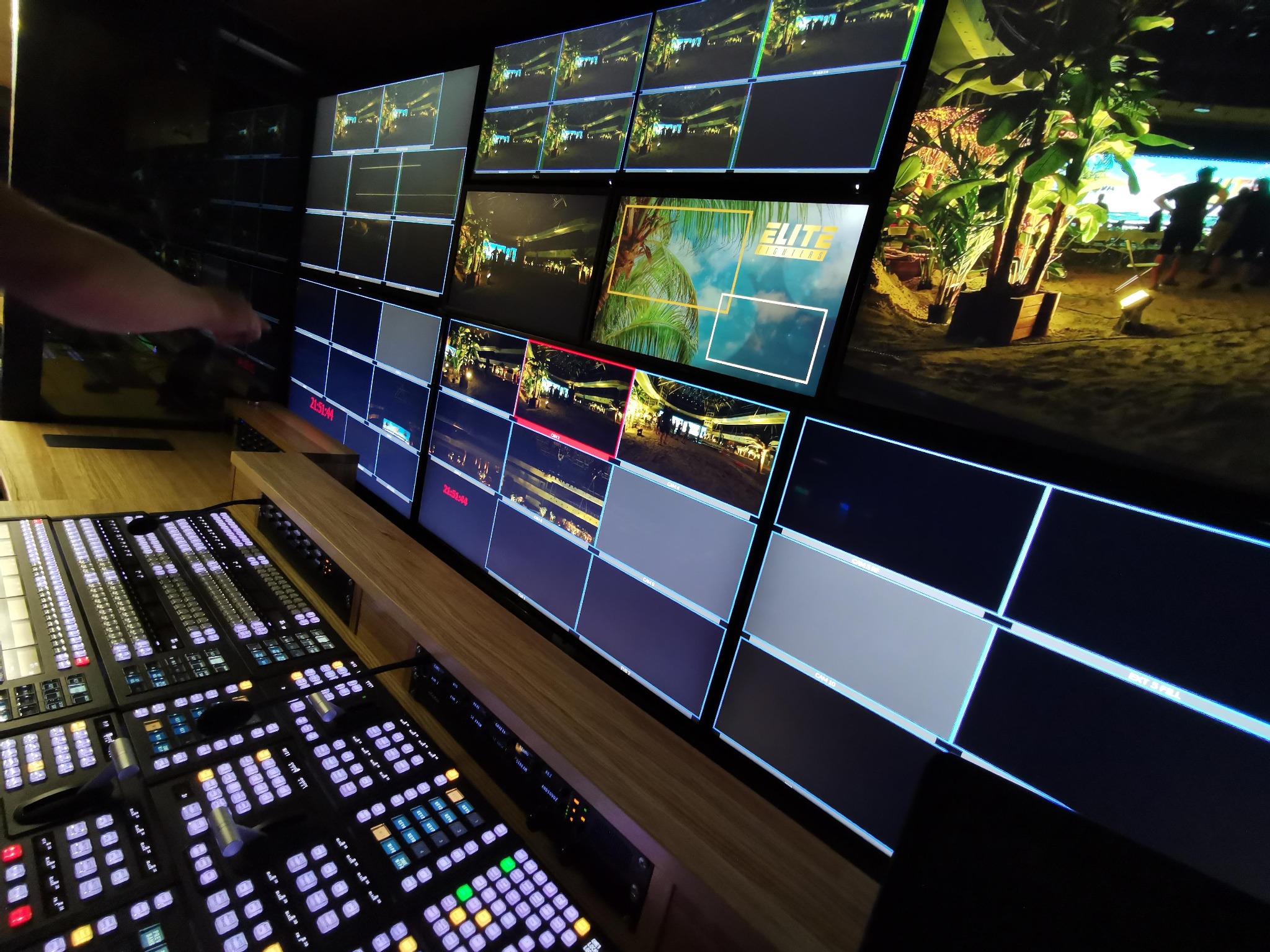 Stanowisko reżyserskie z mikserem wideo i ścianą monitorów wyświetlających obraz z kamer podczas realizacji wydarzenia plenerowego z palmami i oświetleniem.