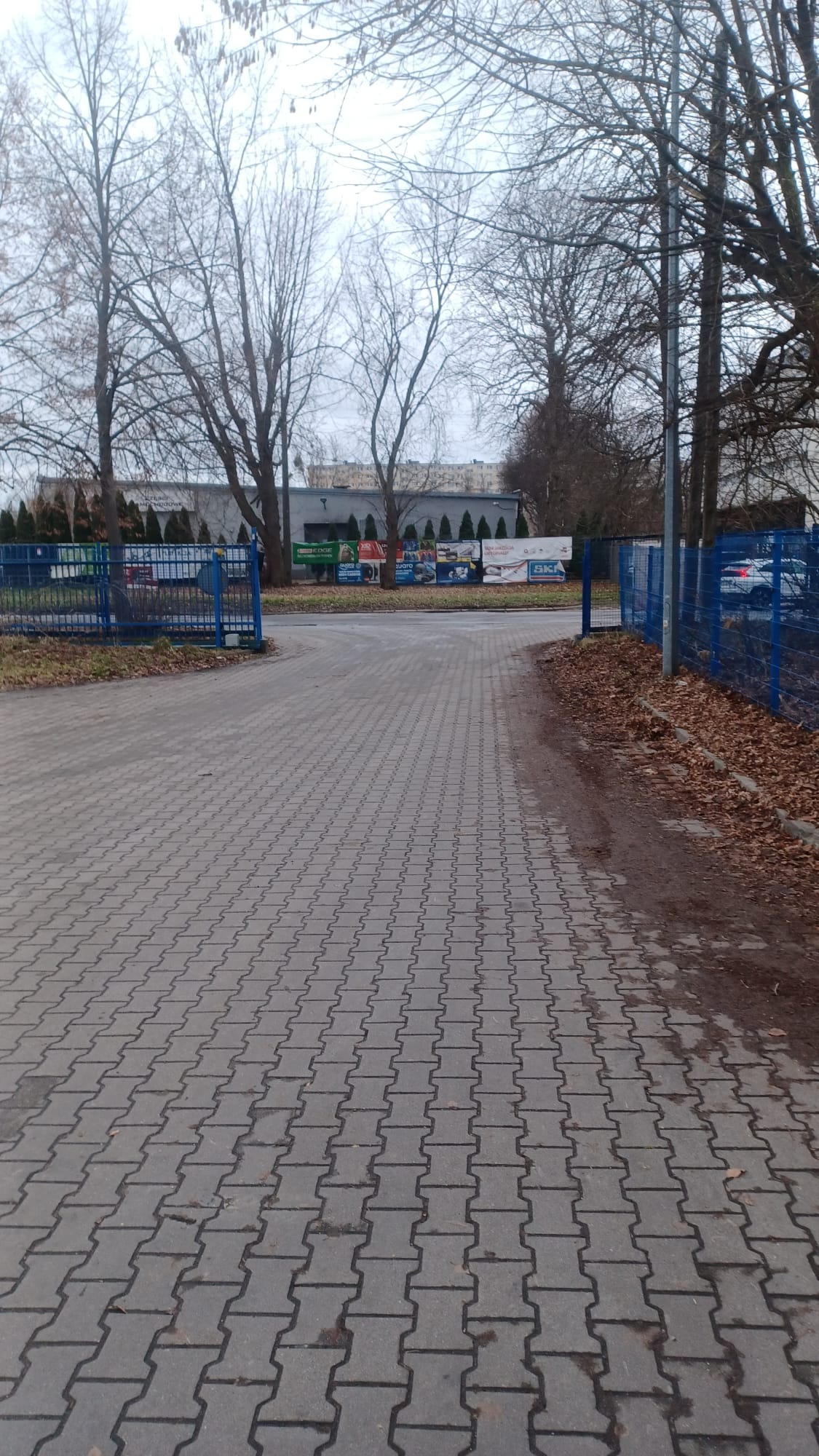Widok z perspektywy pieszego na brukowaną drogę wjazdową z niebieską bramą, prowadzącą do magazynu z widocznymi banerami reklamowymi i zaparkowanym samochodem w tle, w pochmurny dzień.