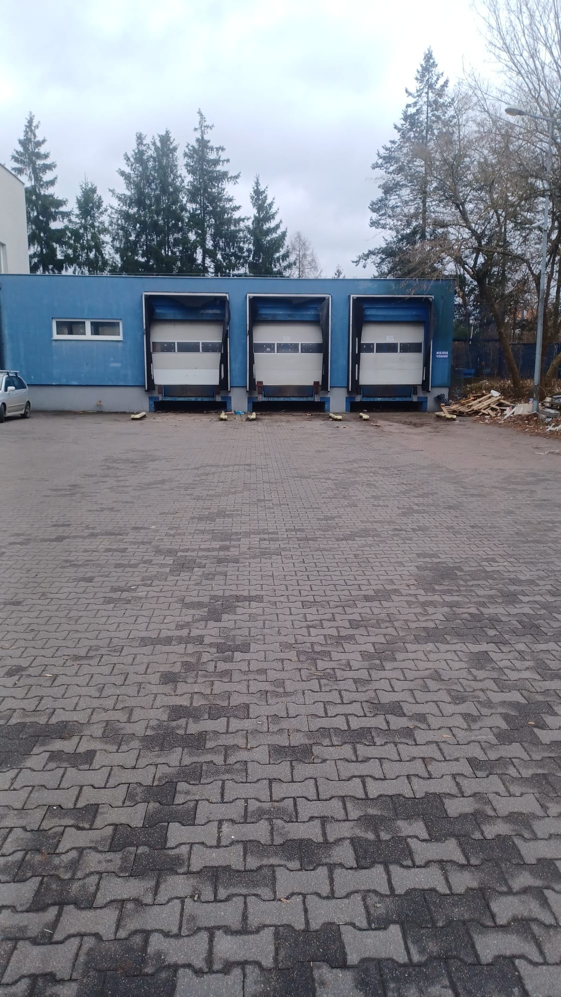 Niebieski budynek magazynowy z trzema bramami załadunkowymi, kostka brukowa na placu, drzewa w tle, pochmurne niebo.