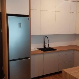 Jakub&Robson - Nowoczesna kuchnia z białymi górnymi szafkami, szarymi dolnymi szafkami i drewnianym blatem, z czarną baterią i zlewozmywakiem, oraz srebrną lodówką w zabudowie.