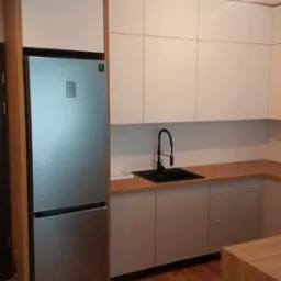 Nowoczesna kuchnia z białymi górnymi szafkami, szarymi dolnymi szafkami i drewnianym blatem, z czarną baterią i zlewozmywakiem, oraz srebrną lodówką w zabudowie.