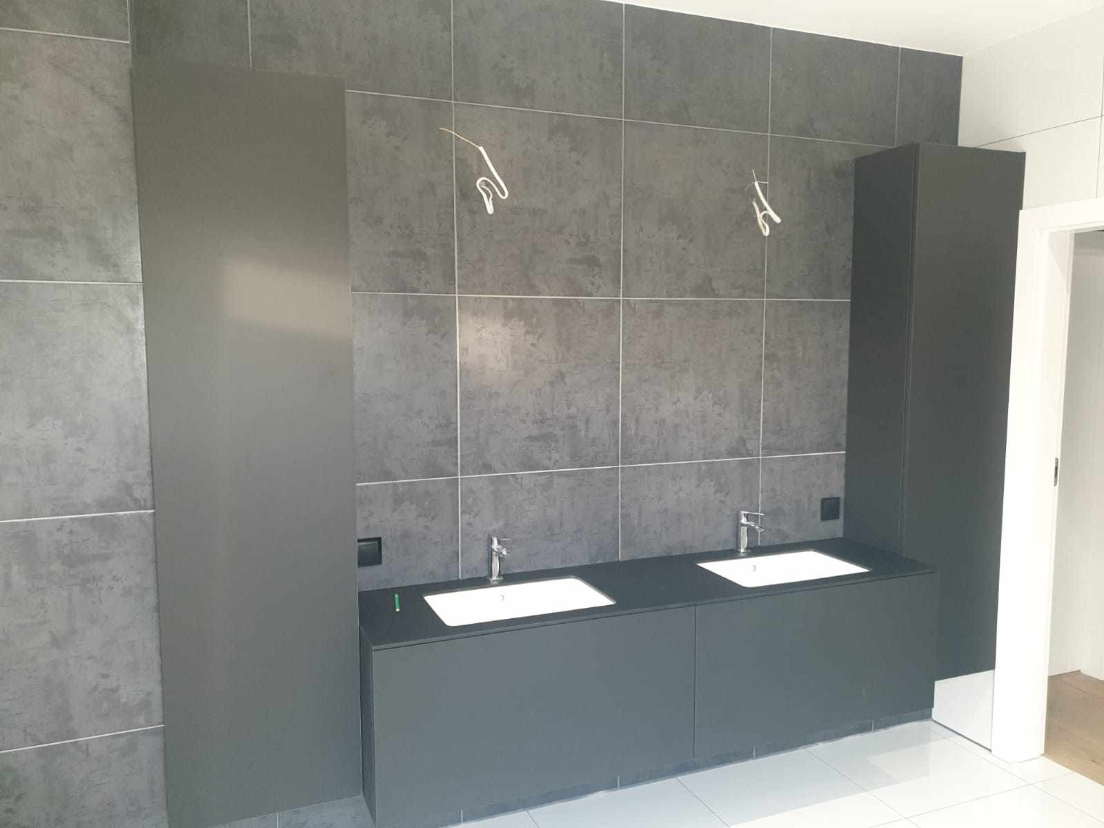 Nowoczesna łazienka z dwoma umywalkami wpuszczonymi w ciemnoszary blat, pod którym znajdują się szafki. Po bokach blatu stoją wysokie, ciemnoszare szafki. Na ścianie szare płytki imitujące beton.