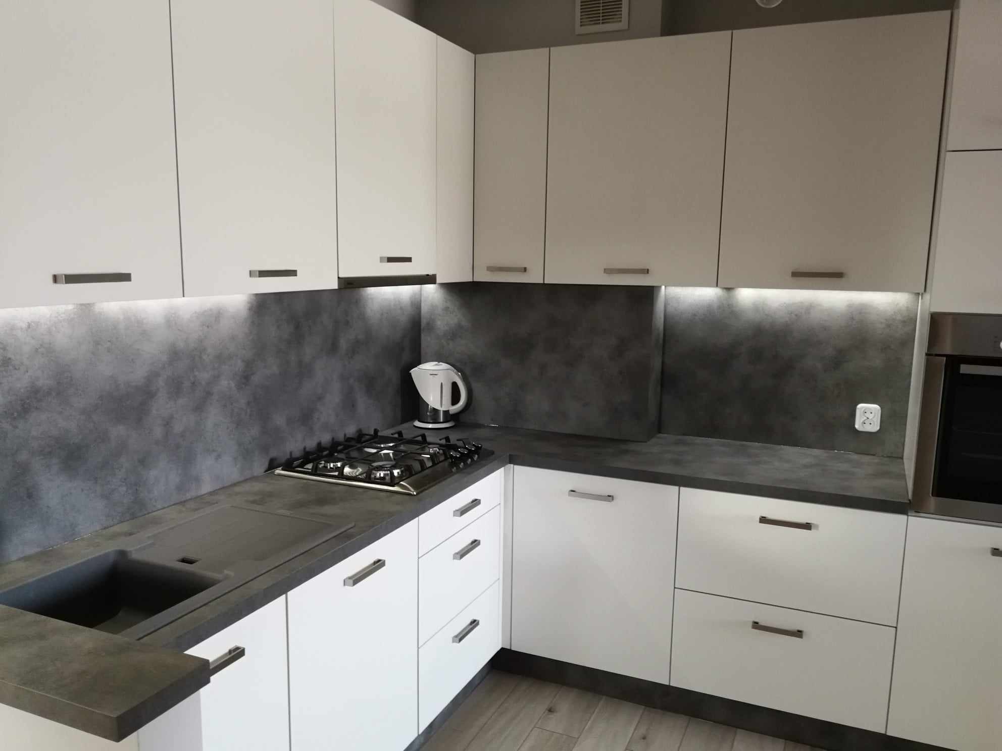 Nowoczesna, narożna zabudowa kuchenna z białymi frontami, szarym blatem i podświetleniem LED pod szafkami wiszącymi. Na blacie znajduje się płyta gazowa i czajnik.