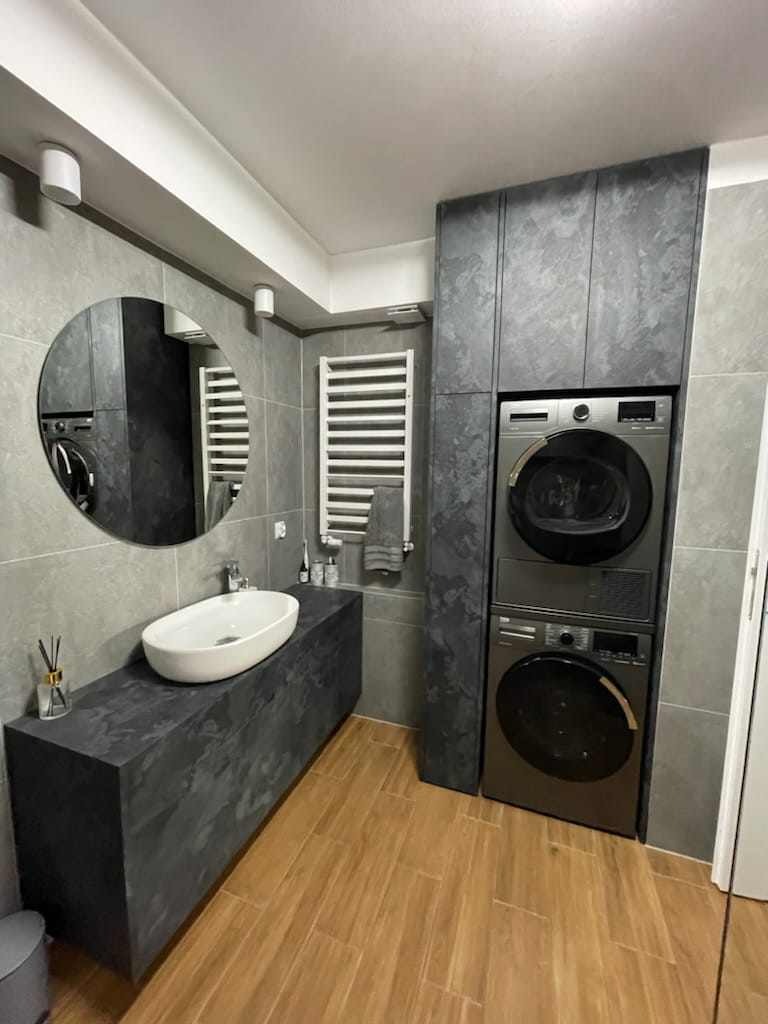 Nowoczesna łazienka z szarymi płytkami, drewnopodobną podłogą, umywalką nablatową na blacie z ciemnego kamienia, okrągłym lustrem, grzejnikiem drabinkowym i pralko-suszarką w zabudowie sięgającej...