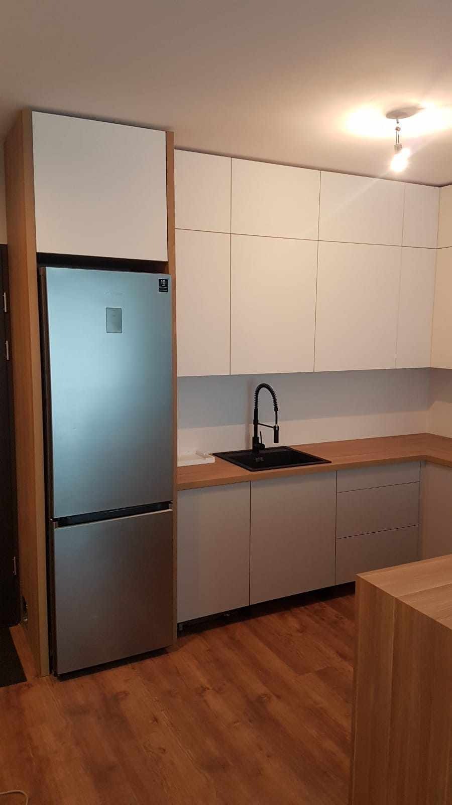 Nowoczesna kuchnia z białymi górnymi szafkami, szarymi dolnymi szafkami i drewnianym blatem, z czarną baterią i zlewozmywakiem, oraz srebrną lodówką w zabudowie.