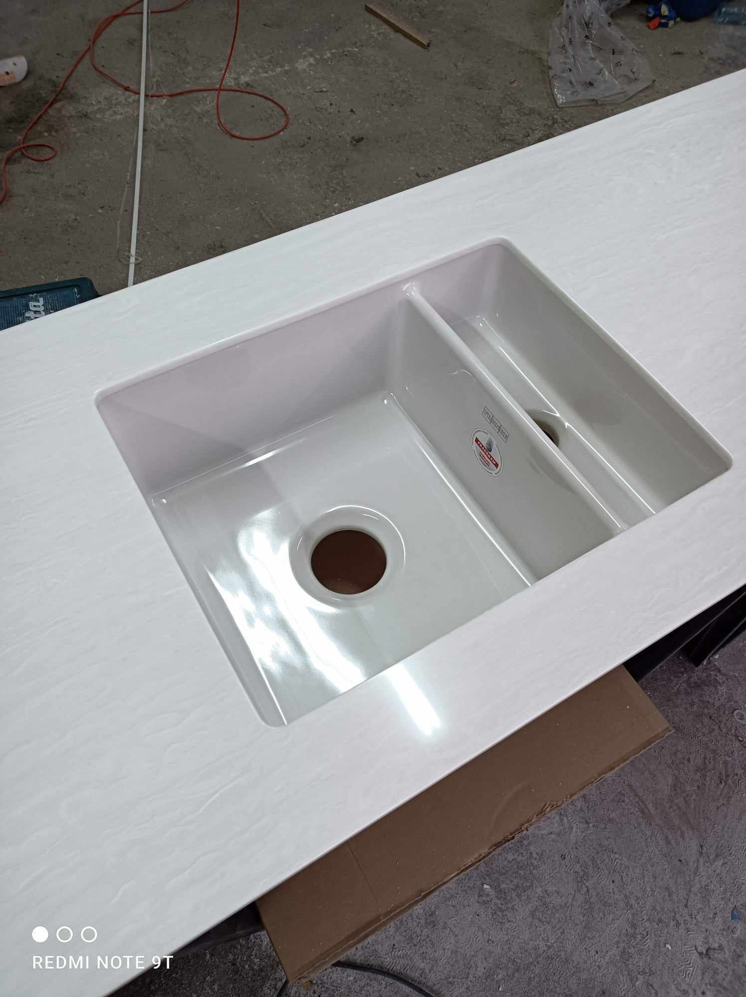 Biały blat kuchenny z wbudowanym, dwukomorowym zlewozmywakiem ceramicznym z okrągłym odpływem, widoczny fragment etykiety producenta.