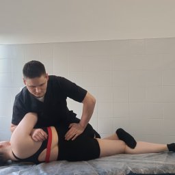 Dynamis Fizjoterapia & Rehabilitacja - Mężczyzna w czarnym uniformie uciska plecy pacjentki leżącej na stole do masażu w jasnym gabinecie z oknem