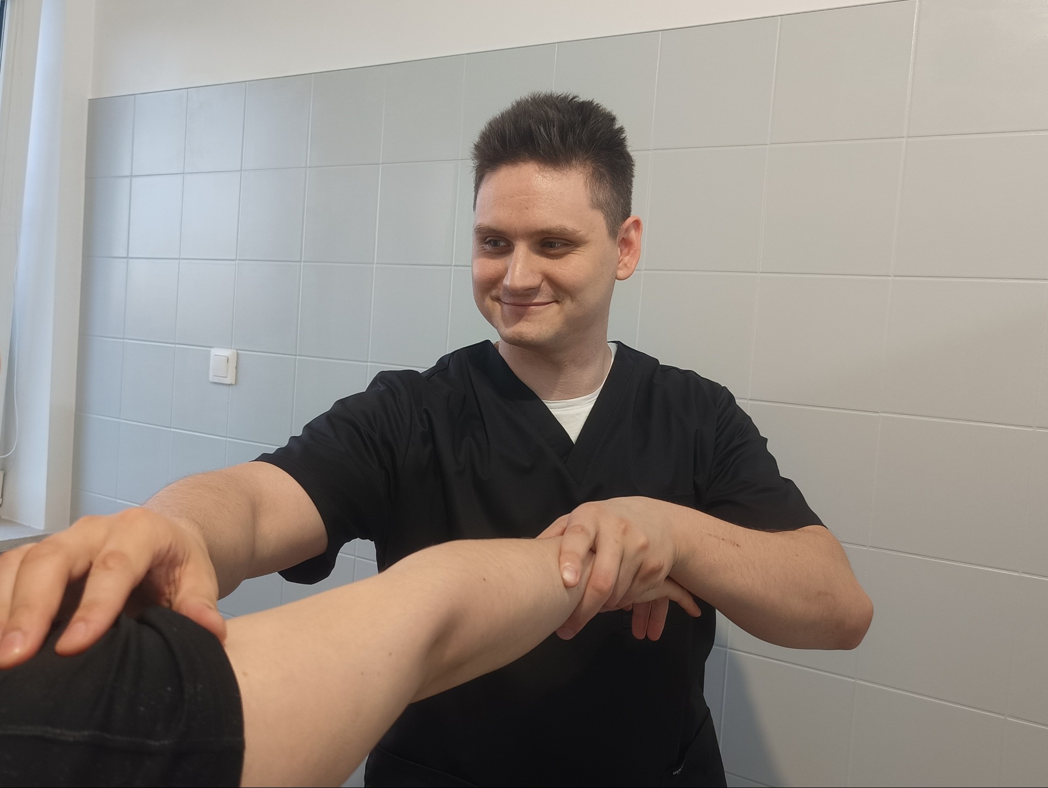 Fizjoterapeuta w czarnym uniformie delikatnie bada ramię pacjenta w gabinecie z jasnymi kafelkami w tle.