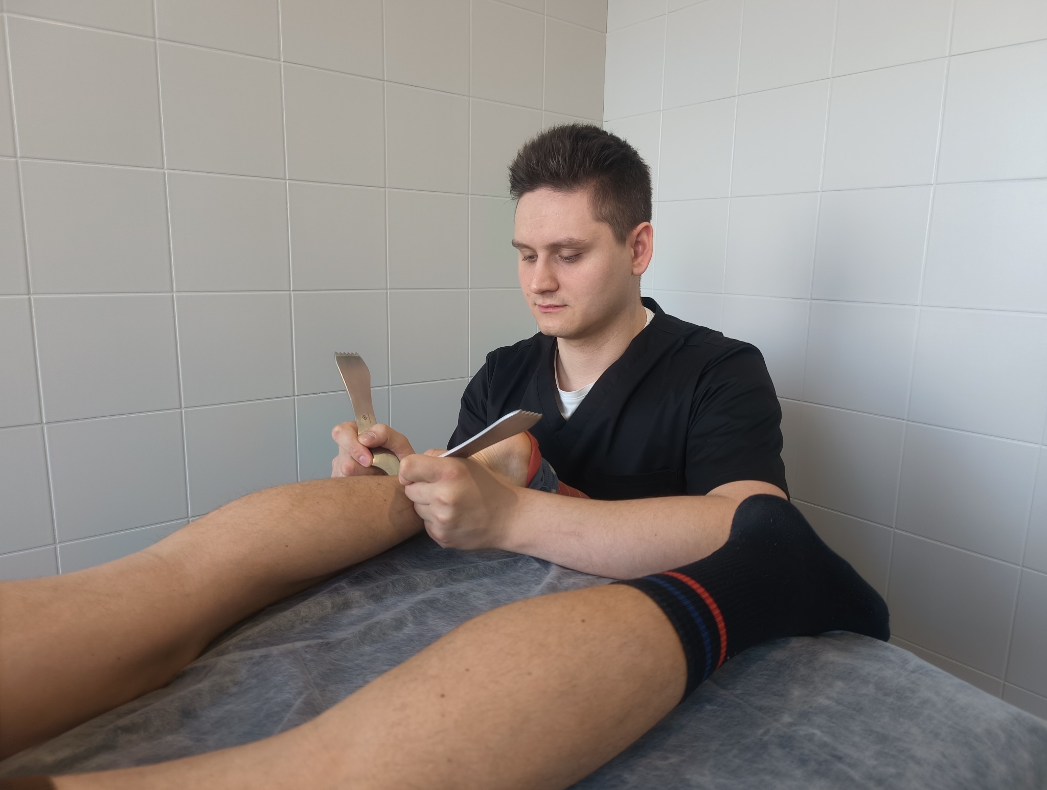 Mężczyzna w czarnym uniformie wykonuje zabieg na nodze pacjenta za pomocą metalowych narzędzi z ząbkowanymi krawędziami na tle białych płytek.