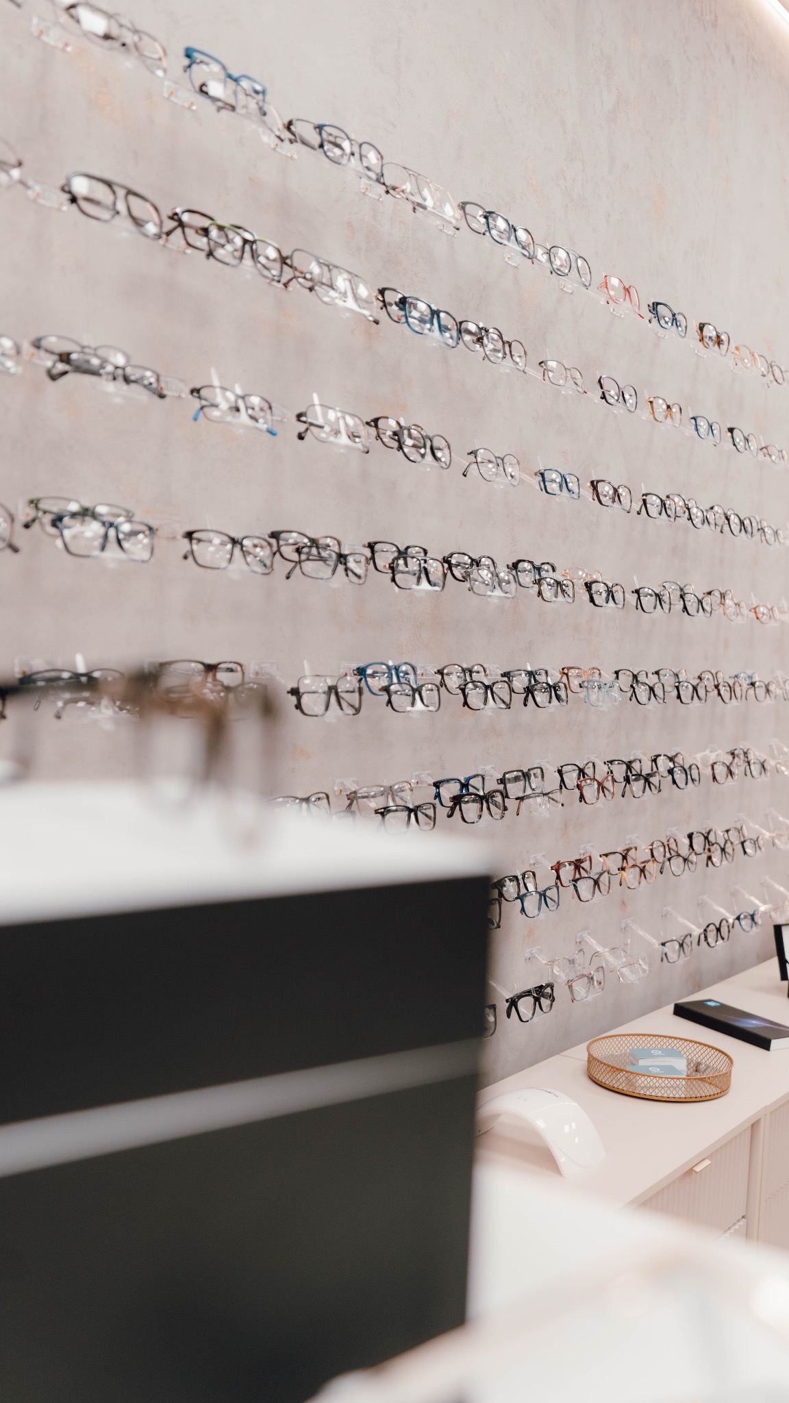 Ekspozycja okularów korekcyjnych i przeciwsłonecznych na ścianie w salonie optycznym, z elementami wyposażenia na pierwszym planie.