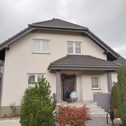 Hydro - Dwupiętrowy dom z beżową elewacją w trakcie prac wykończeniowych; widoczny pracownik przygotowujący podłoże przy wejściu, zabezpieczone folią drzwi, szare kolumny i schody wejściowe, oraz zadbany...