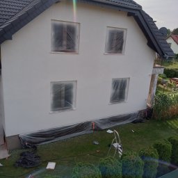 Hydro - Dom jednorodzinny z nową, jasną elewacją. Okna zabezpieczone folią, a u podstawy budynku rozłożona folia ochronna. W tle widoczny ogród i fragment sąsiedniego domu.