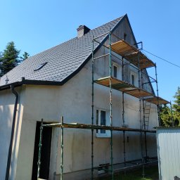 Hydro - Dom jednorodzinny w trakcie prac elewacyjnych, widoczne rusztowanie, surowy tynk i częściowo pomalowany dach, błękitne niebo w tle.