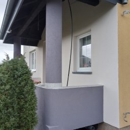 Hydro - Wykończona elewacja domu z szarymi kolumnami i podbitką, widoczne okno z białymi ramami, fragment ogrodu z tujami i fragment żółtej ściany.