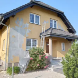 Dwupiętrowy dom z żółtą elewacją w trakcie przygotowań do prac, widoczne odspojenia starej farby, aluminiowa drabina oparta o ścianę, zabezpieczona folią roślina, kostka brukowa na podjeździe.