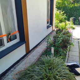 Hydro - Elewacja budynku z oknem zabezpieczonym folią malarską, świeżo pomalowane obramowanie okna w kolorze antracytowym, widoczne drewniane elementy dekoracyjne na elewacji, otoczenie budynku...