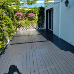 Szara deska tarasowa kompozytowa na zewnątrz, z białym pergolą porośniętą zielenią i różowymi kwiatami, przylegająca do białego budynku z czarną lampą zewnętrzną.