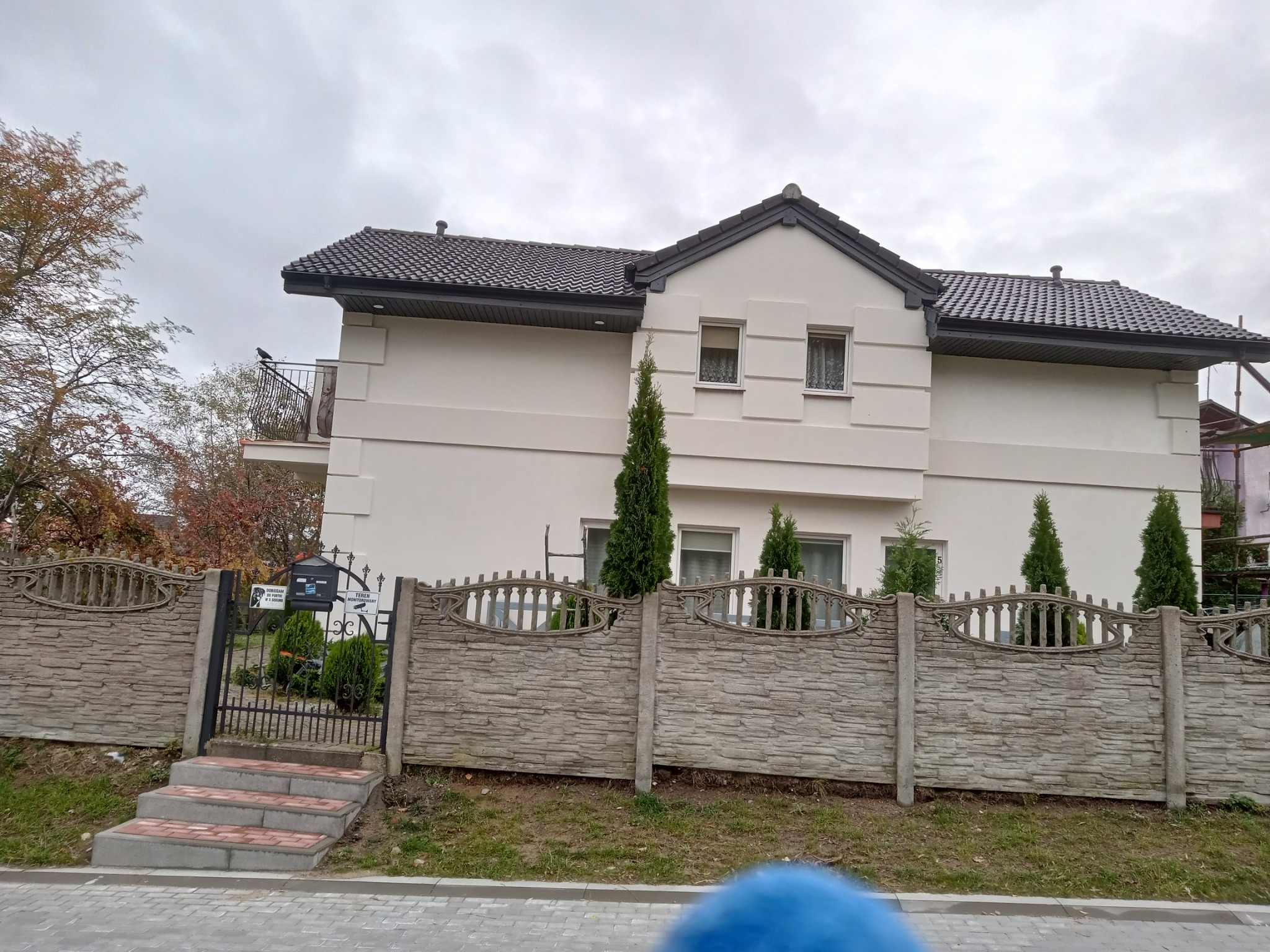 Dwukondygnacyjny dom z jasną elewacją, ciemną dachówką i balkonem, widziany zza kamiennego ogrodzenia z metalową furtką i schodami wejściowymi; pochmurne niebo w tle.