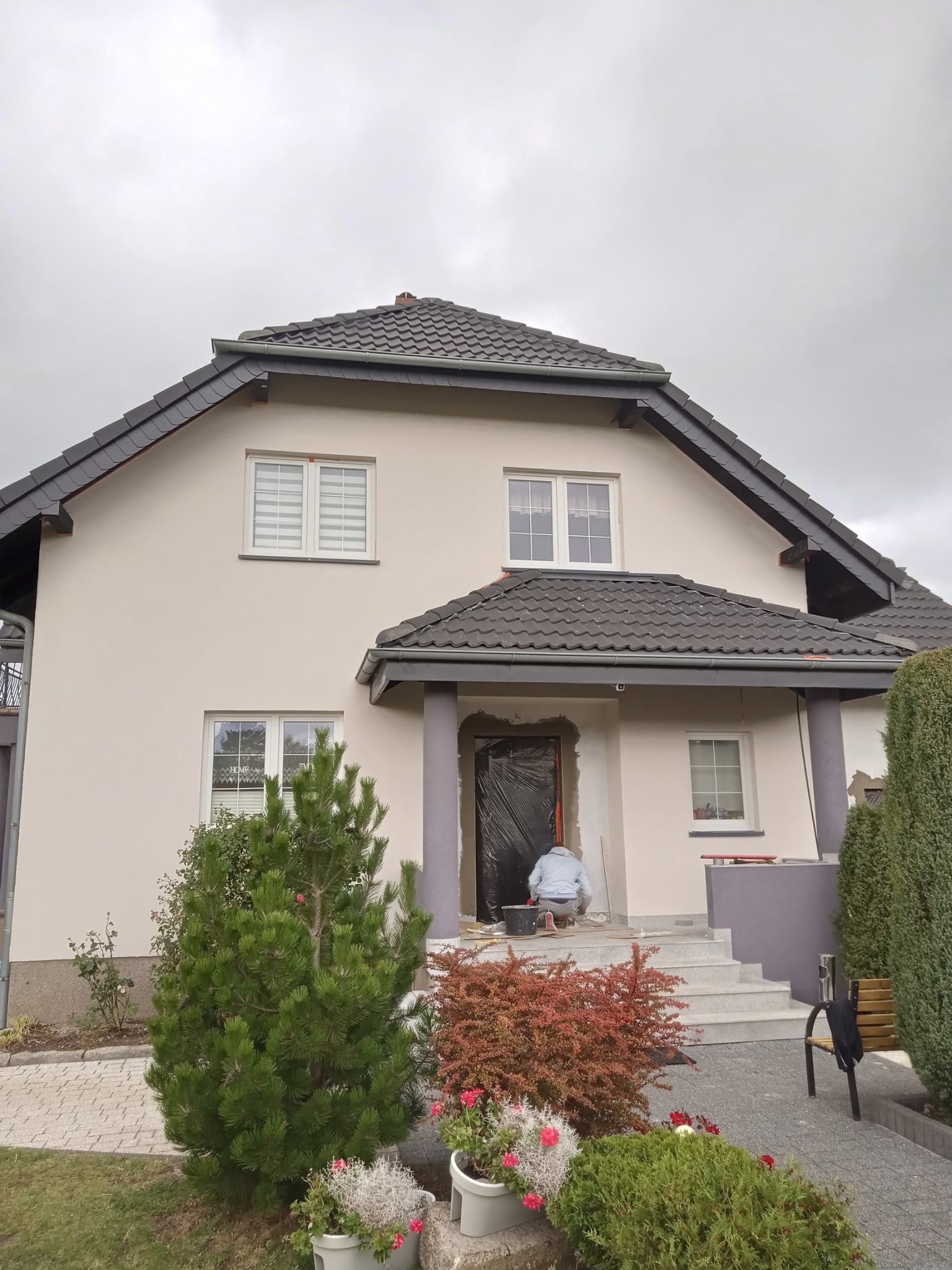 Dwupiętrowy dom z beżową elewacją w trakcie prac wykończeniowych; widoczny pracownik przygotowujący podłoże przy wejściu, zabezpieczone folią drzwi, szare kolumny i schody wejściowe, oraz zadbany...