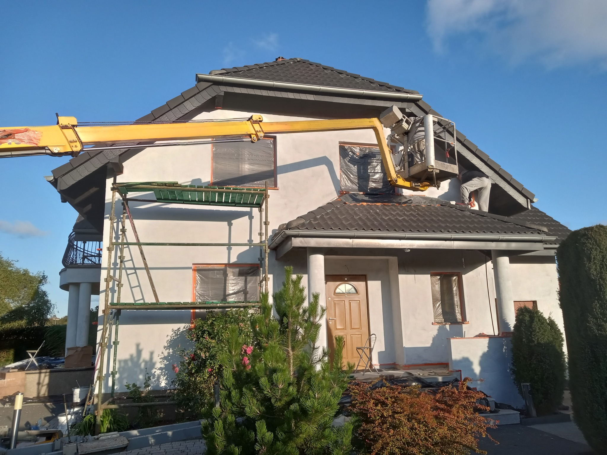 Prace na elewacji domu jednorodzinnego z użyciem podnośnika koszowego i rusztowania, okna zabezpieczone folią, widoczne ślady świeżo nałożonego tynku.