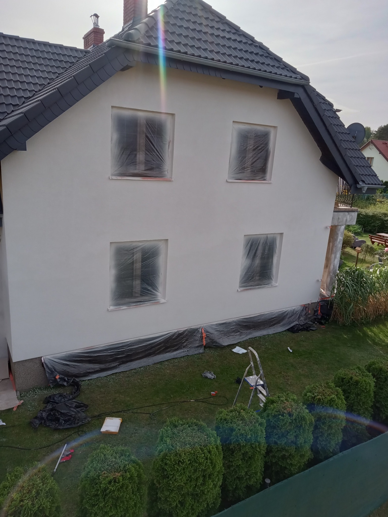 Dom jednorodzinny z nową, jasną elewacją. Okna zabezpieczone folią, a u podstawy budynku rozłożona folia ochronna. W tle widoczny ogród i fragment sąsiedniego domu.