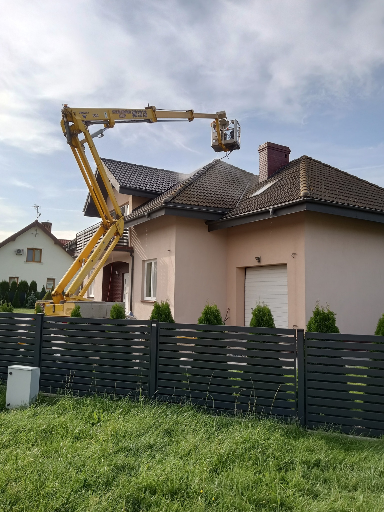 Pracownik na podnośniku koszowym czyści dach domu jednorodzinnego strumieniem wody pod ciśnieniem. W tle widoczne inne budynki i błękitne niebo.