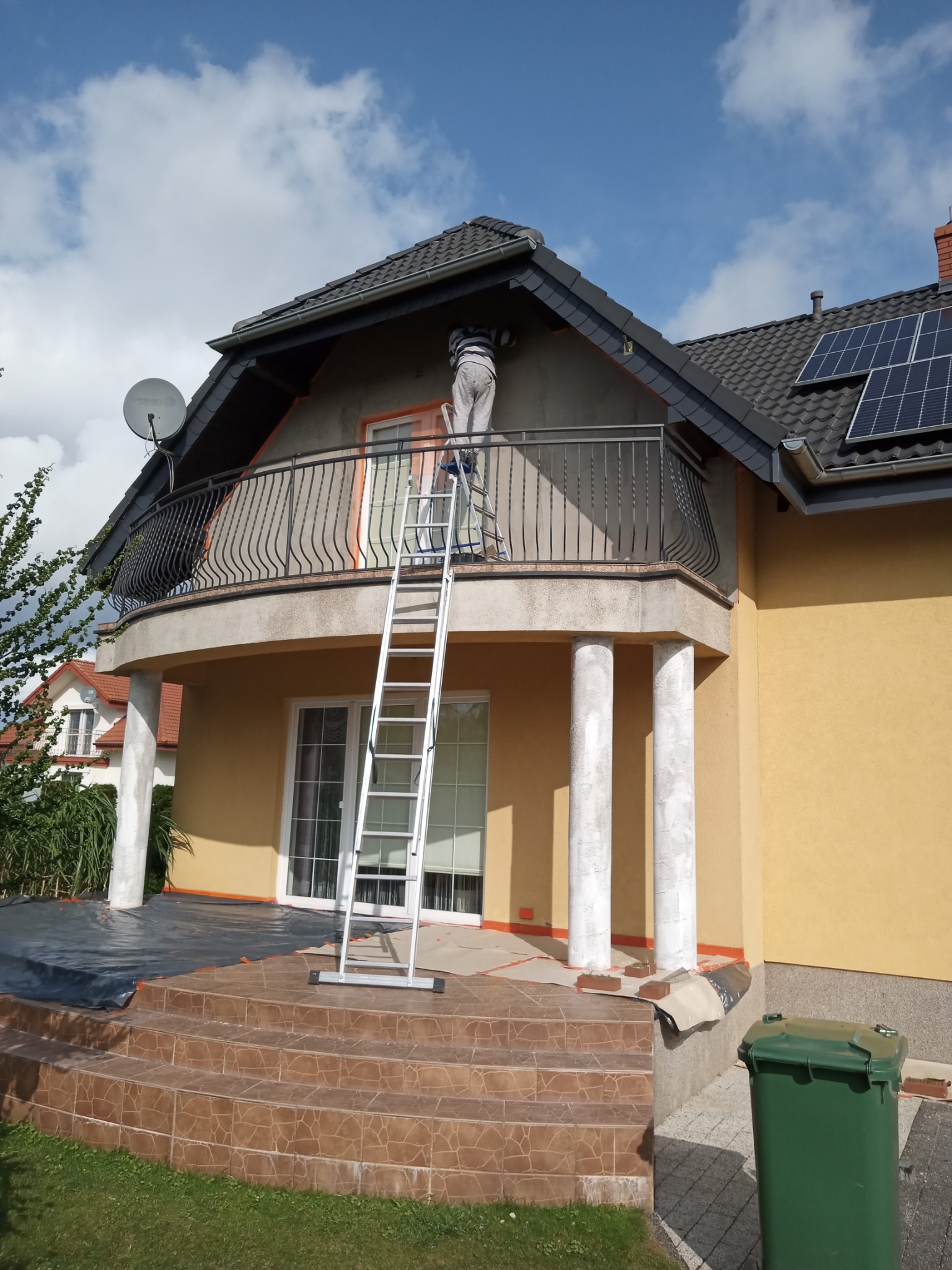 Pracownik na drabinie maluje elewację balkonu domu jednorodzinnego z panelami słonecznymi na dachu, widok z dołu na tle błękitnego nieba.