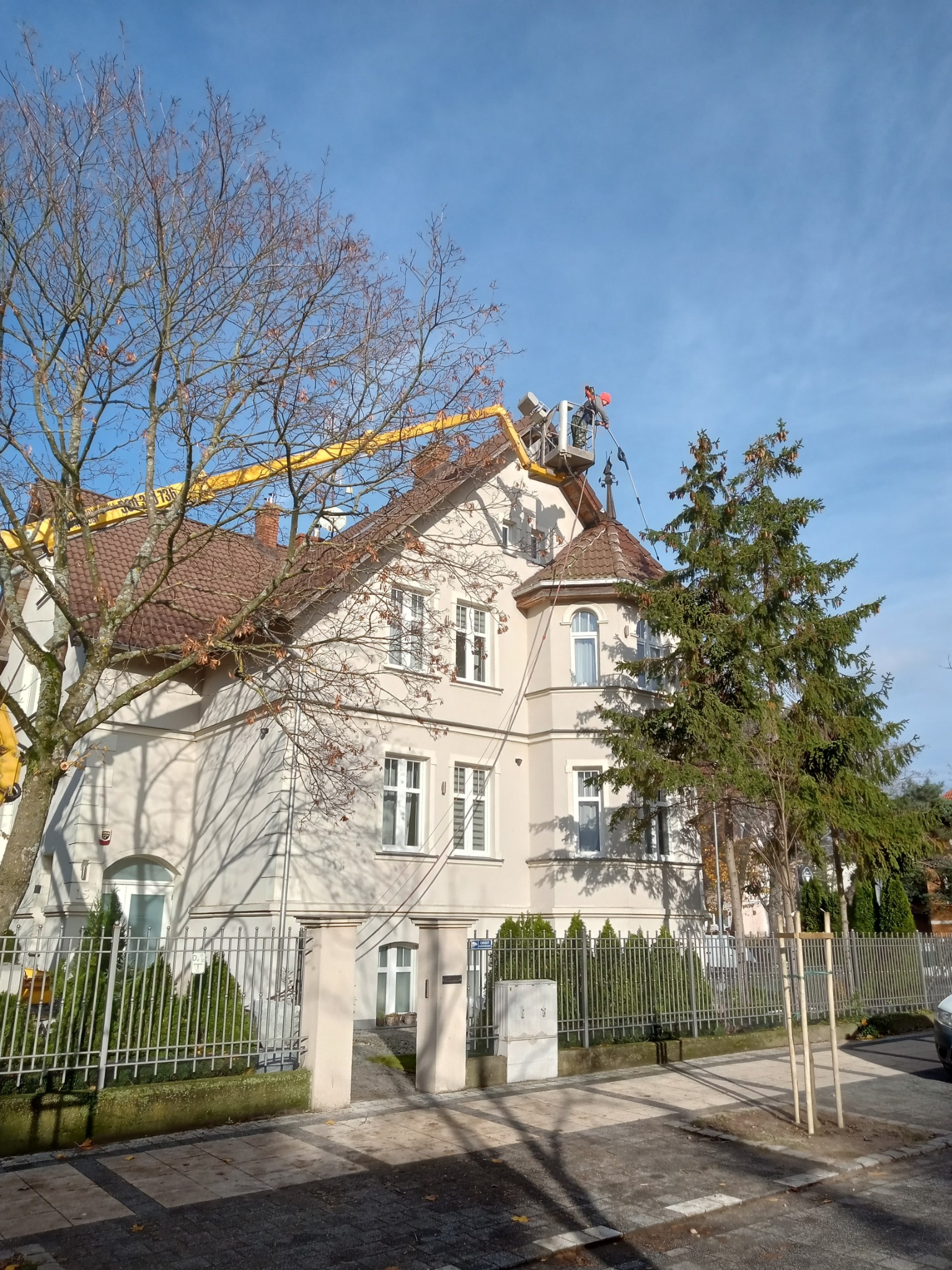 Pracownik na podnośniku koszowym, w trakcie prac na elewacji dwupiętrowego domu z czerwonym dachem i białymi oknami, widok z poziomu ulicy.