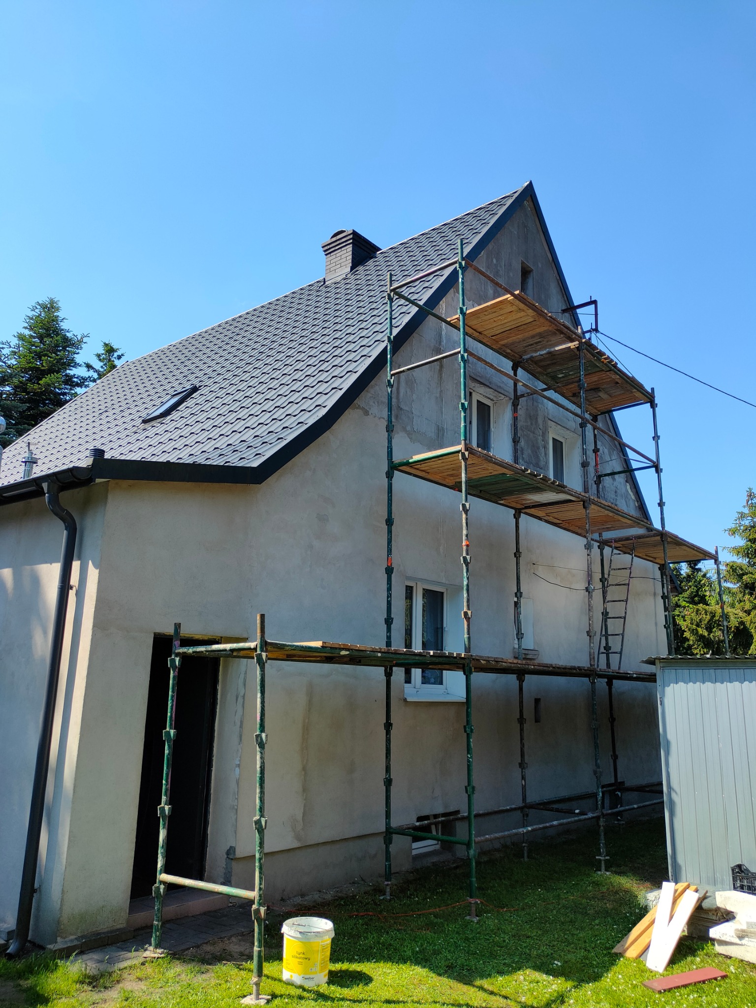 Dom jednorodzinny w trakcie prac elewacyjnych, widoczne rusztowanie, surowy tynk i częściowo pomalowany dach, błękitne niebo w tle.