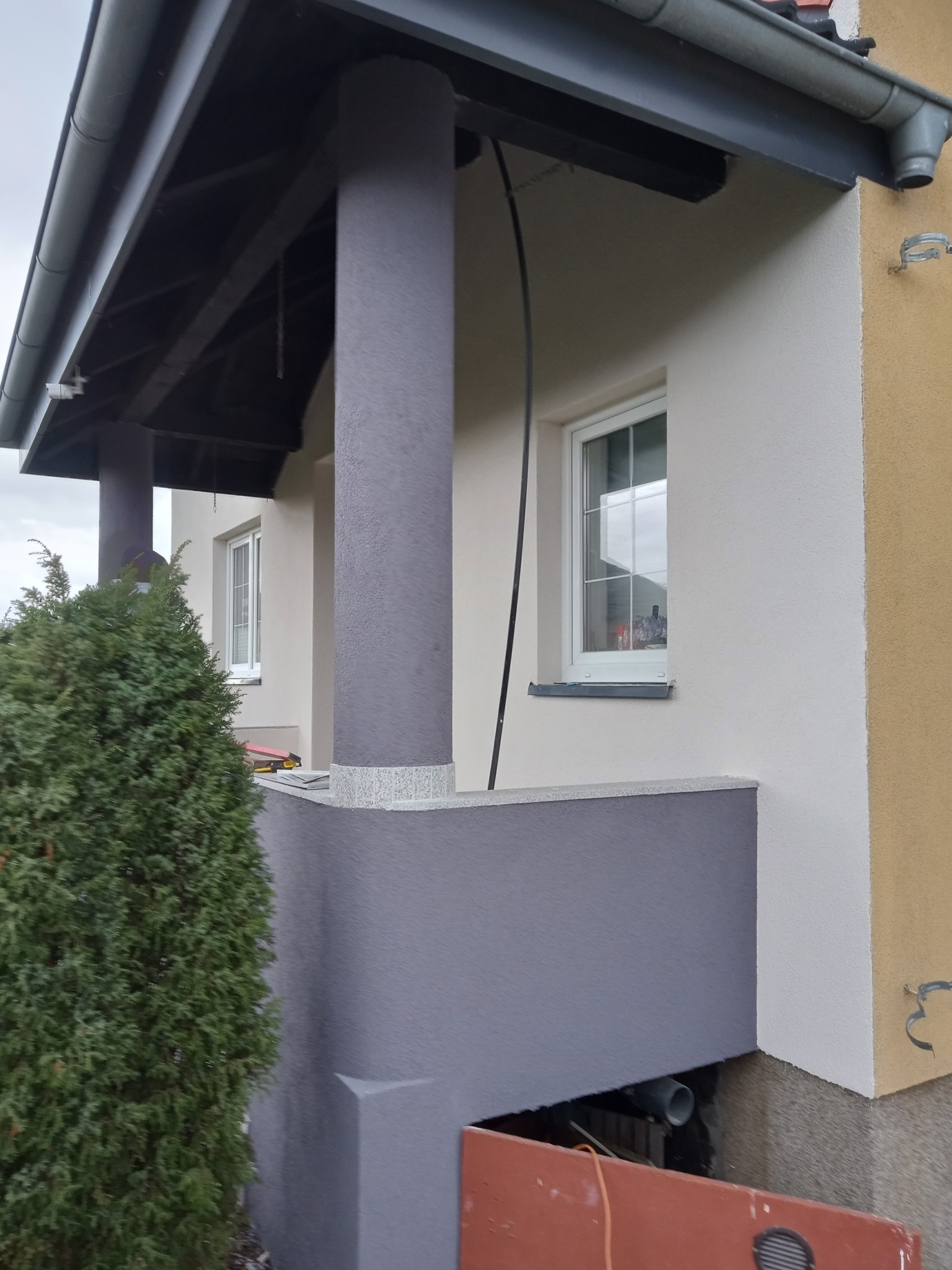 Wykończona elewacja domu z szarymi kolumnami i podbitką, widoczne okno z białymi ramami, fragment ogrodu z tujami i fragment żółtej ściany.