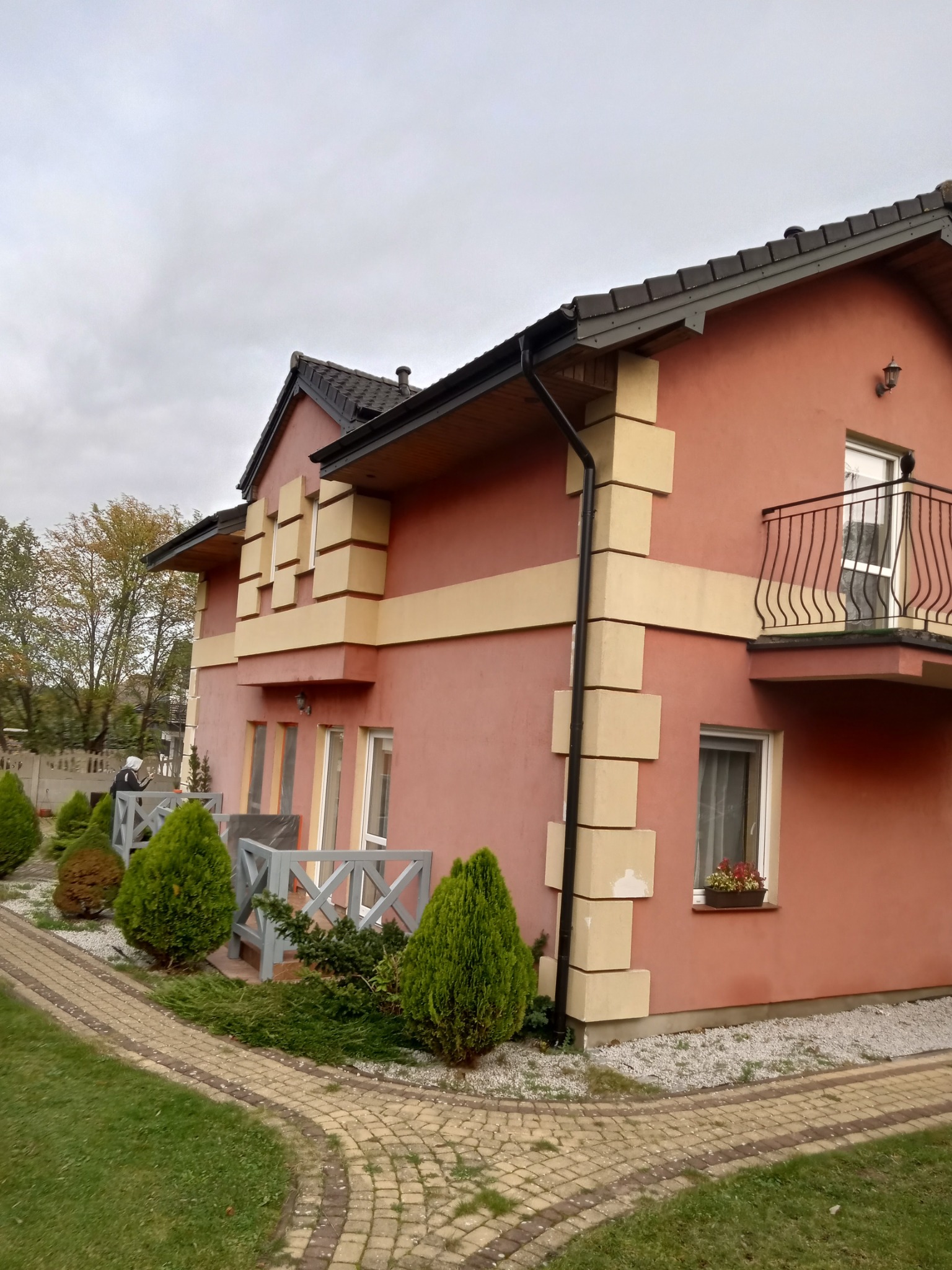 Dwukondygnacyjny dom z pomarańczową elewacją, beżowymi detalami architektonicznymi, balkonem z kutą balustradą i brukowanym chodnikiem.