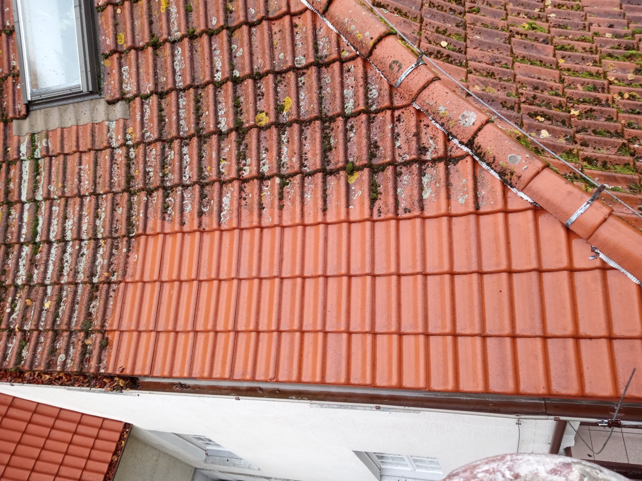 Częściowo wyczyszczony dach z czerwonej dachówki, z widocznymi śladami mchu i porostów na nieoczyszczonej części, okno dachowe w lewym górnym rogu.