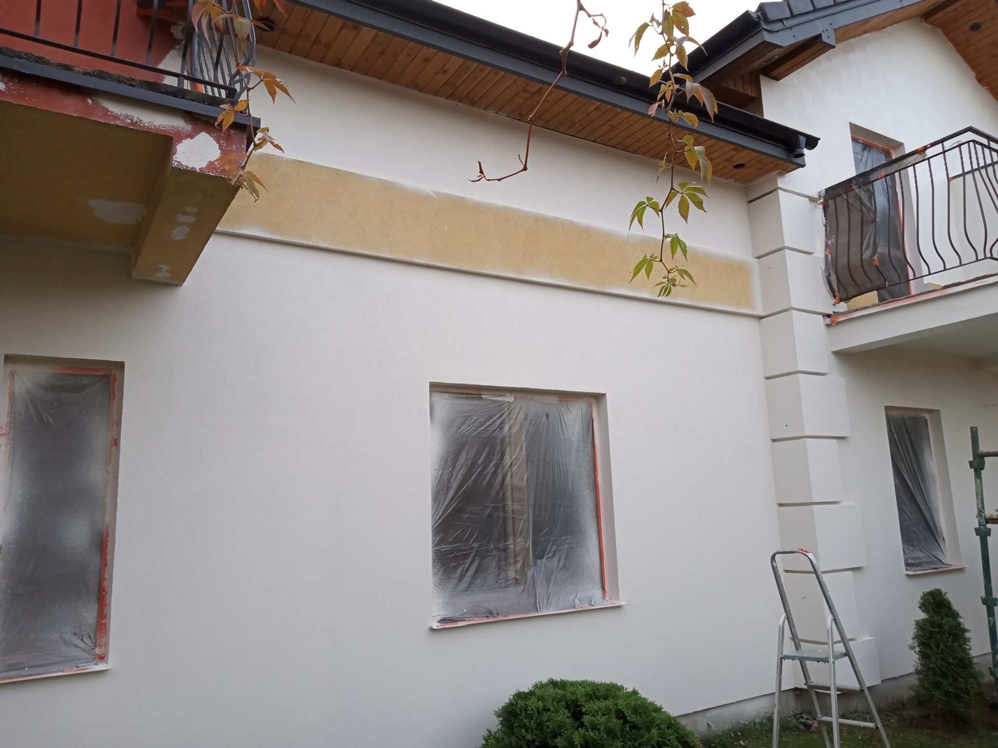 Elewacja budynku w trakcie prac: okna zabezpieczone folią, fragment ściany z nałożonym pasem tynku, drabina oparta o ścianę, krzew przed budynkiem.