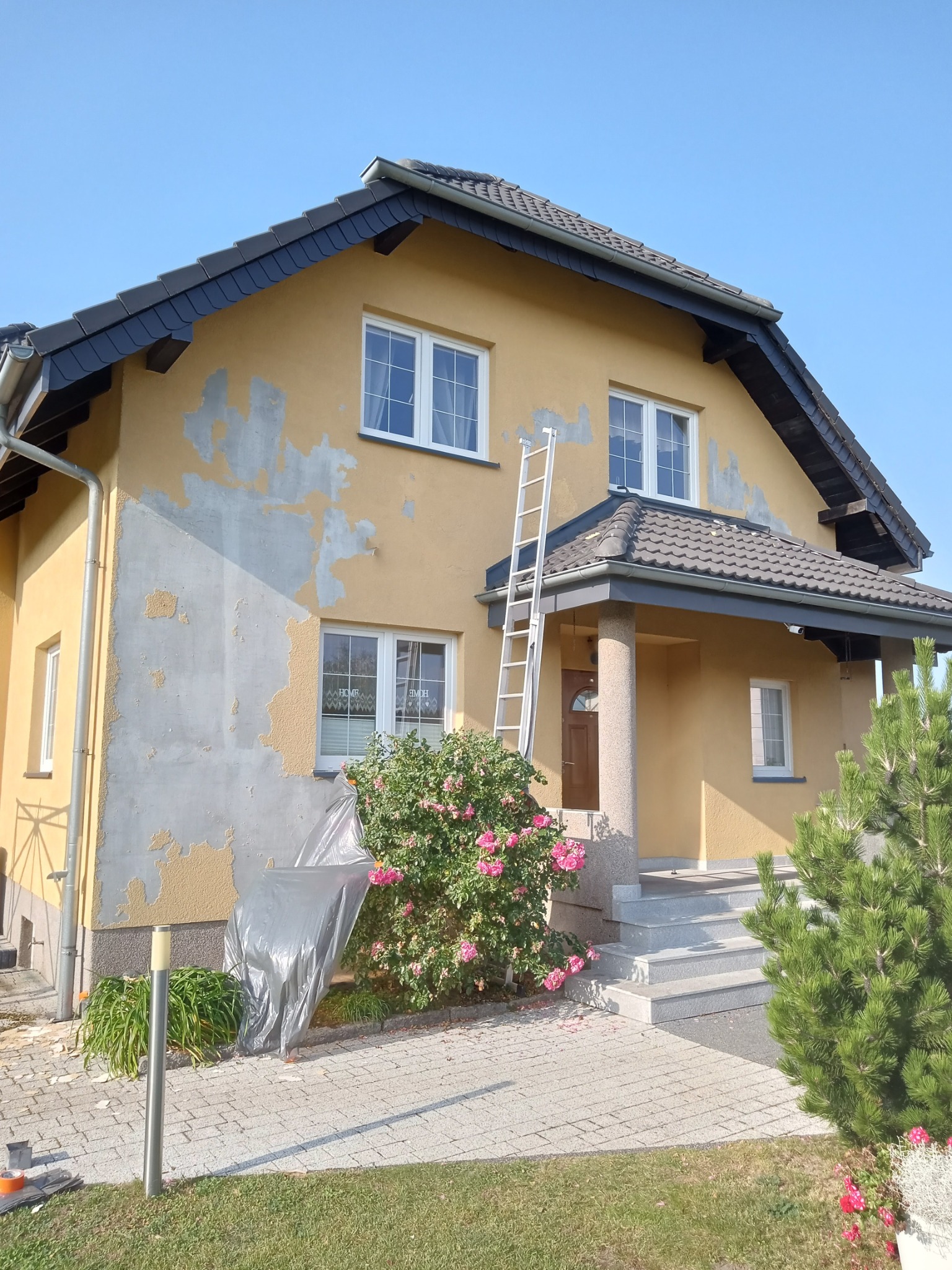 Dwupiętrowy dom z żółtą elewacją w trakcie przygotowań do prac, widoczne odspojenia starej farby, aluminiowa drabina oparta o ścianę, zabezpieczona folią roślina, kostka brukowa na podjeździe.