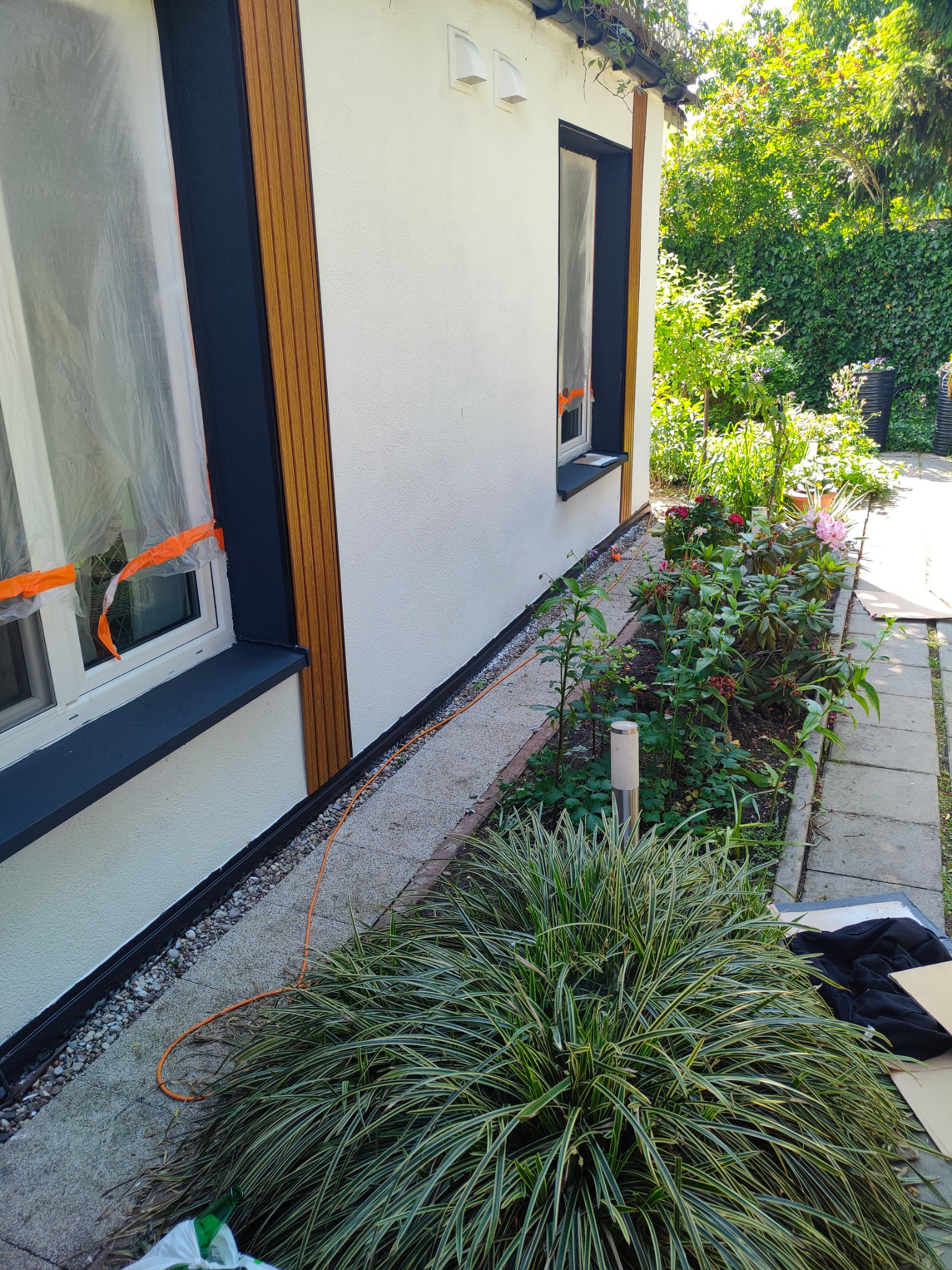 Elewacja budynku z oknem zabezpieczonym folią malarską, świeżo pomalowane obramowanie okna w kolorze antracytowym, widoczne drewniane elementy dekoracyjne na elewacji, otoczenie budynku...