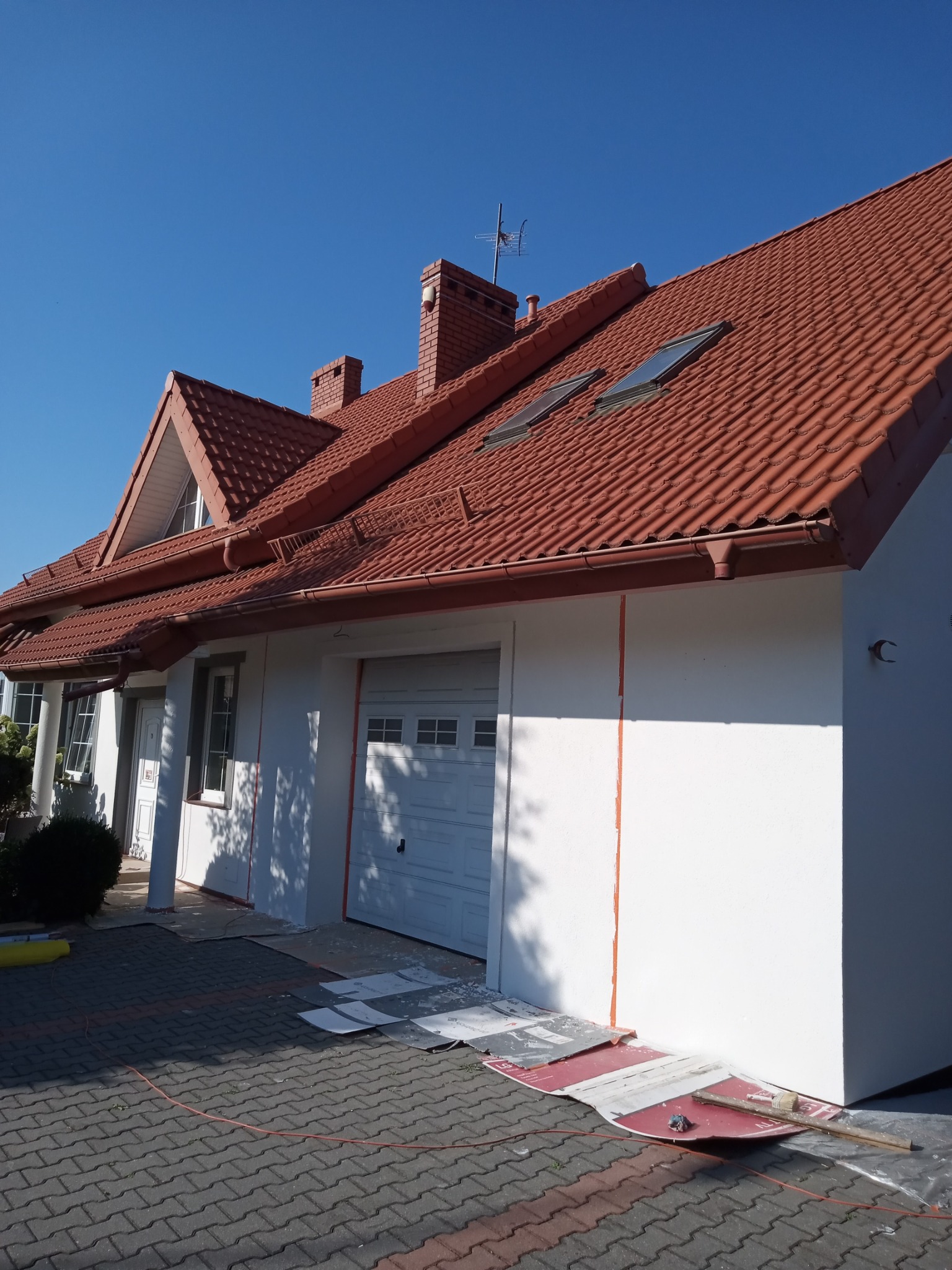Dom jednorodzinny z czerwoną dachówką po częściowym pomalowaniu elewacji na biało, widoczne zabezpieczenia na chodniku i resztki farby.