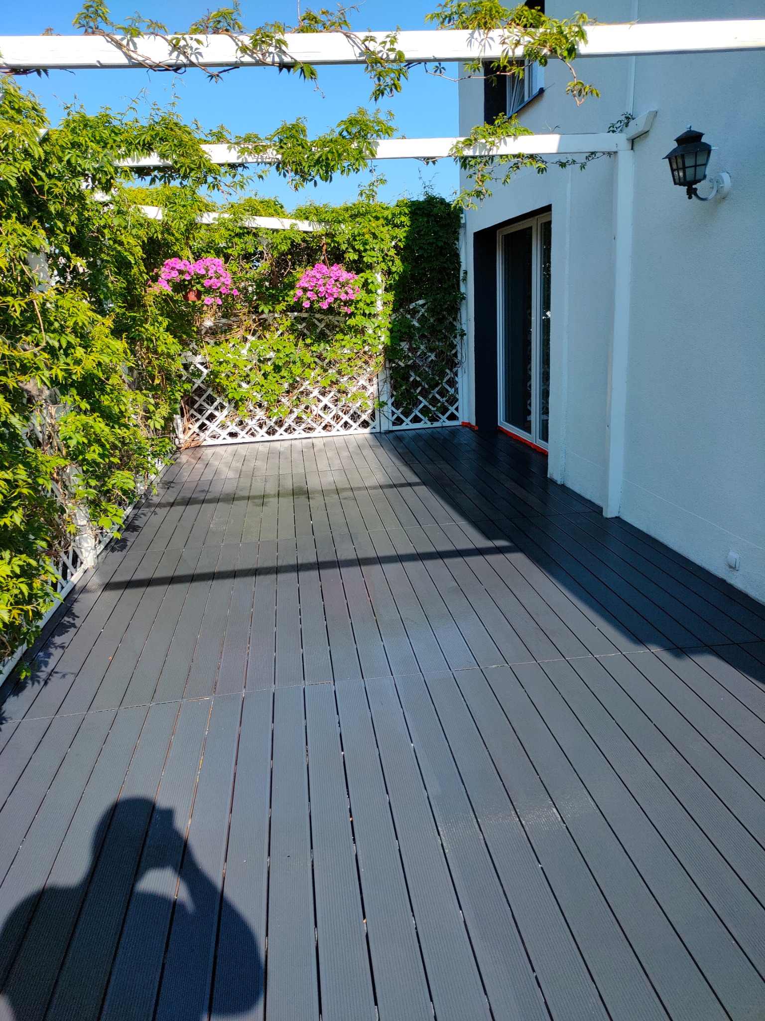 Szara deska tarasowa kompozytowa na zewnątrz, z białym pergolą porośniętą zielenią i różowymi kwiatami, przylegająca do białego budynku z czarną lampą zewnętrzną.
