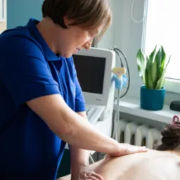 Kobieta w niebieskiej koszulce z logo wykonuje masaż pleców pacjentowi w gabinecie z aparaturą do fizykoterapii i rośliną doniczkową na parapecie.