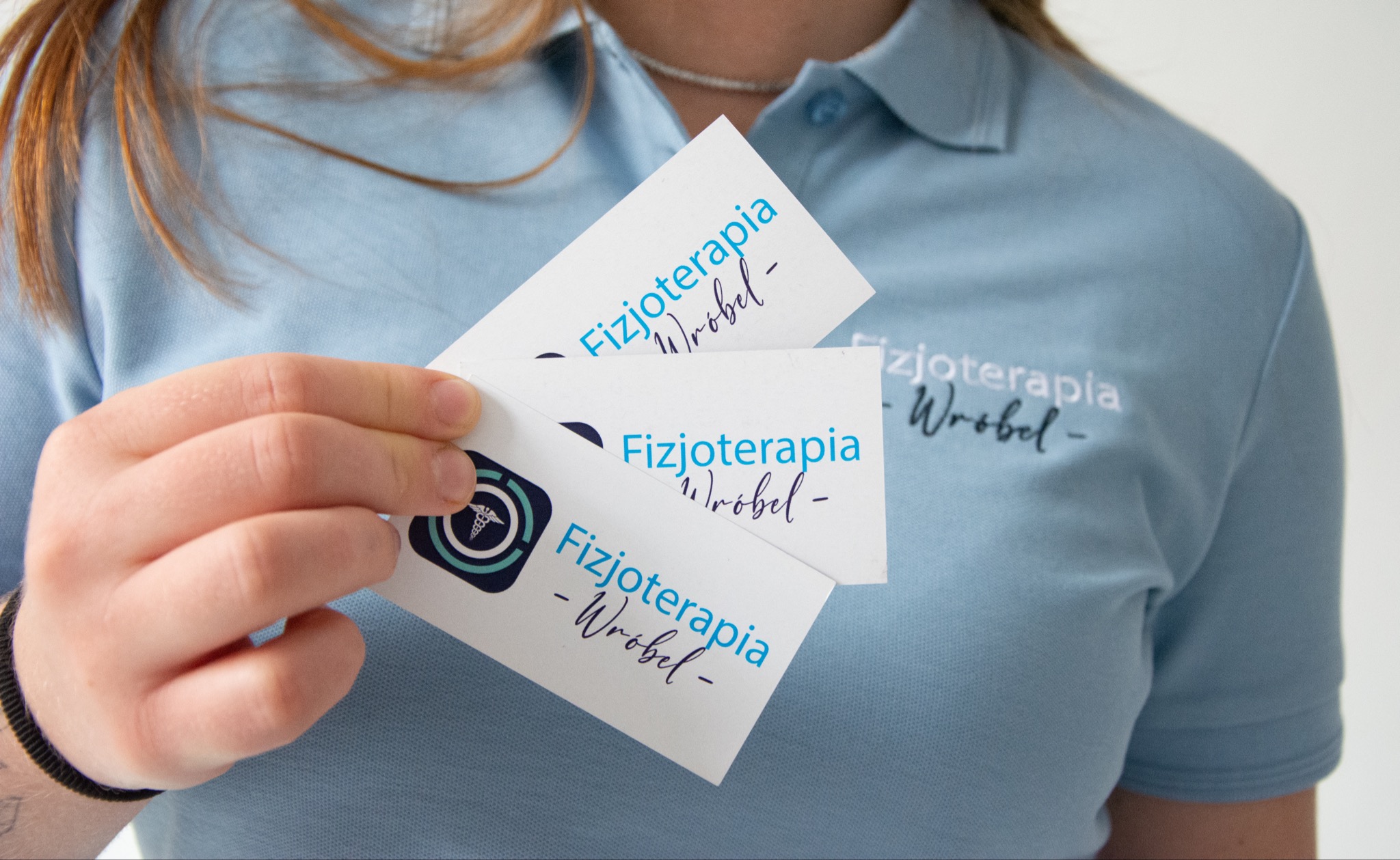Osoba w błękitnej koszulce polo prezentuje wizytówki z napisem 'Fizjoterapia Wróbel' i logo z symbolem Eskulapa w okręgu.