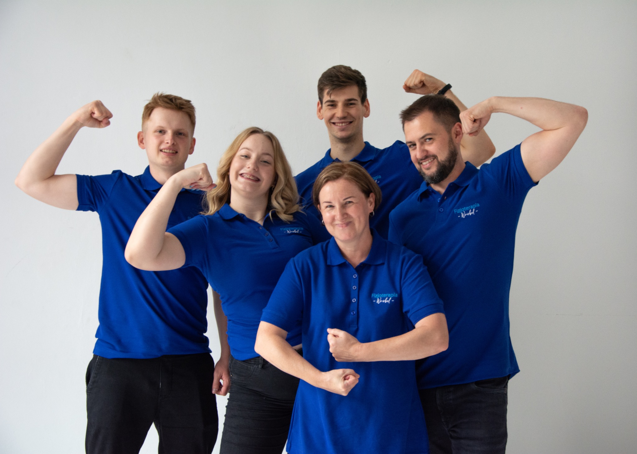 Grupa pięciu uśmiechniętych osób w niebieskich polówkach z logo, prezentujących napięte bicepsy na jasnym tle.